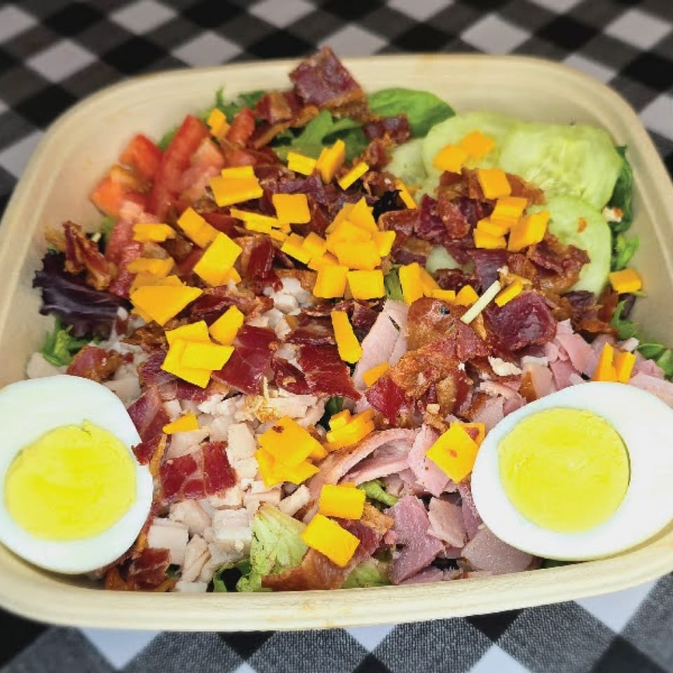A deli salad with lettuce, tomatoes, cucumbers, bacon, ham, turkey, cheddar cheese, and hard-boiled eggs.